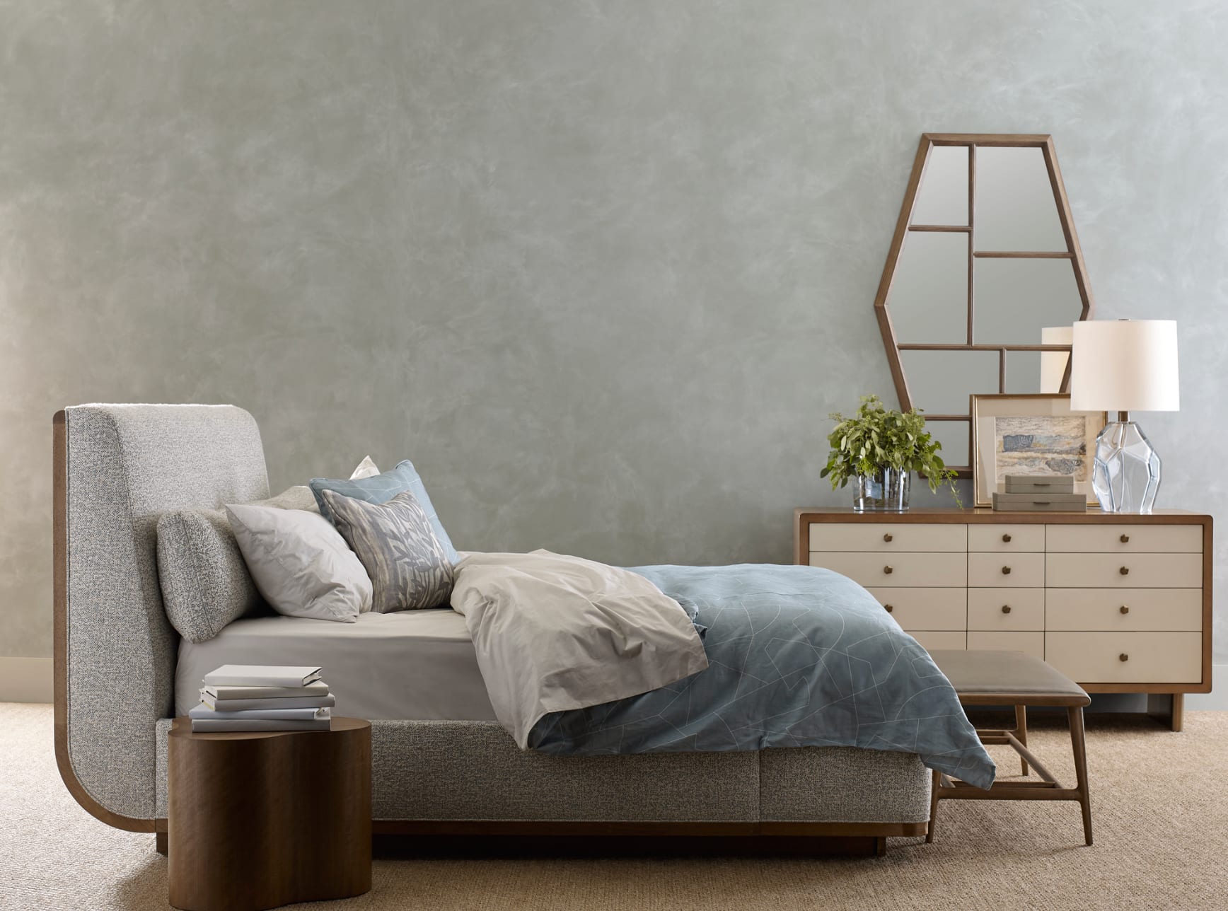 A bedroom in soft sage limewash — a curved bouclé headboard, walnut bench and chest, faceted mirror catching low light