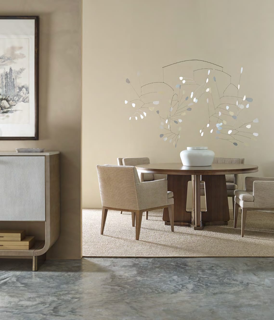 A quiet dining room — round walnut pedestal table, linen armchairs and a kinetic mobile against a plaster wall