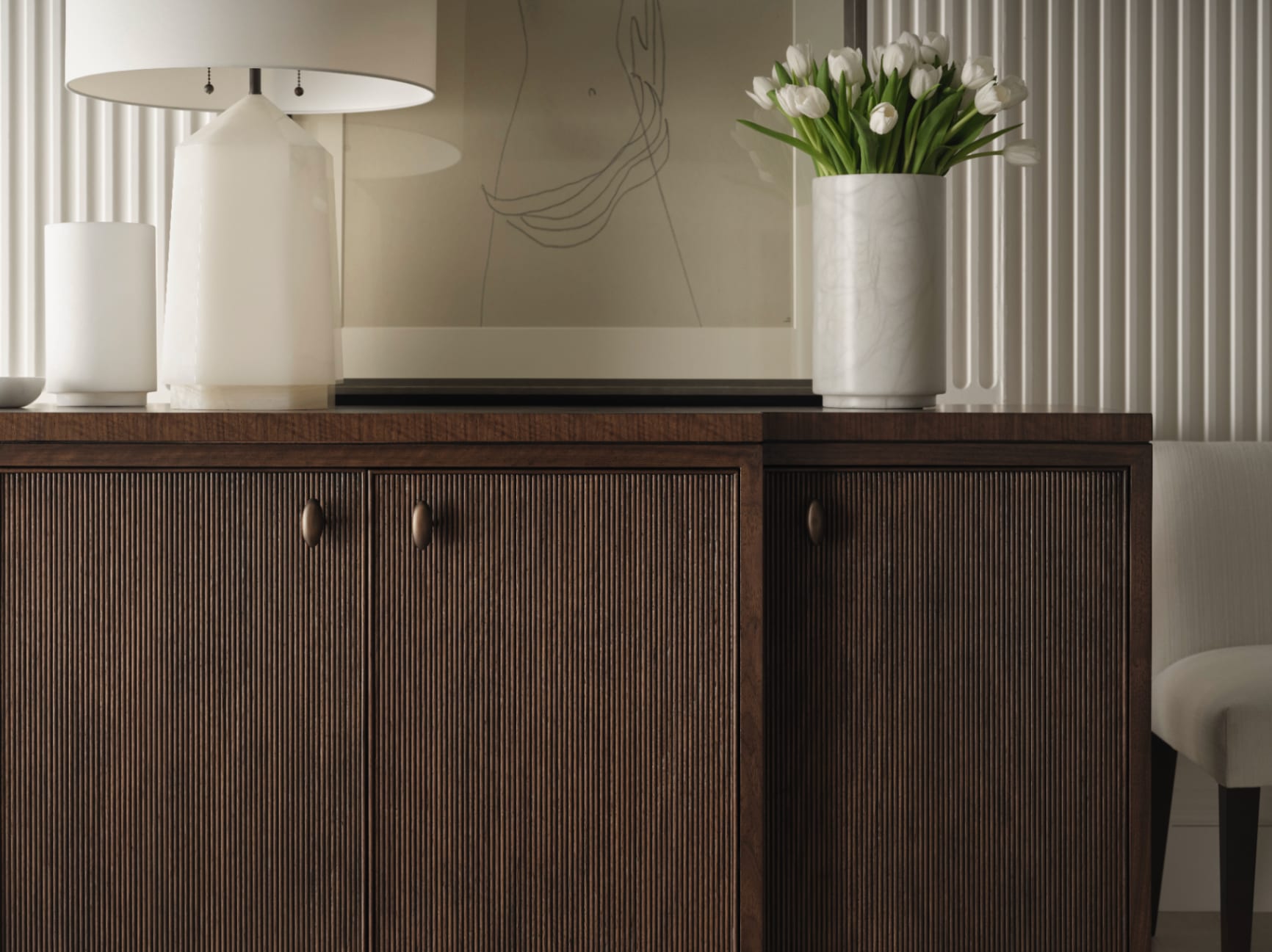 A close detail of a reeded walnut sideboard with antique bronze pulls — alabaster lamp, marble vase of white tulips, fluted plaster wall behind