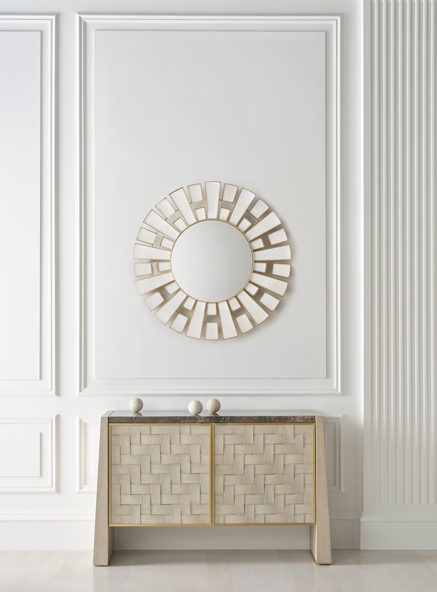 A sunburst mirror in pale gesso and brass above a woven oak console with a marble top, set against panelled plaster