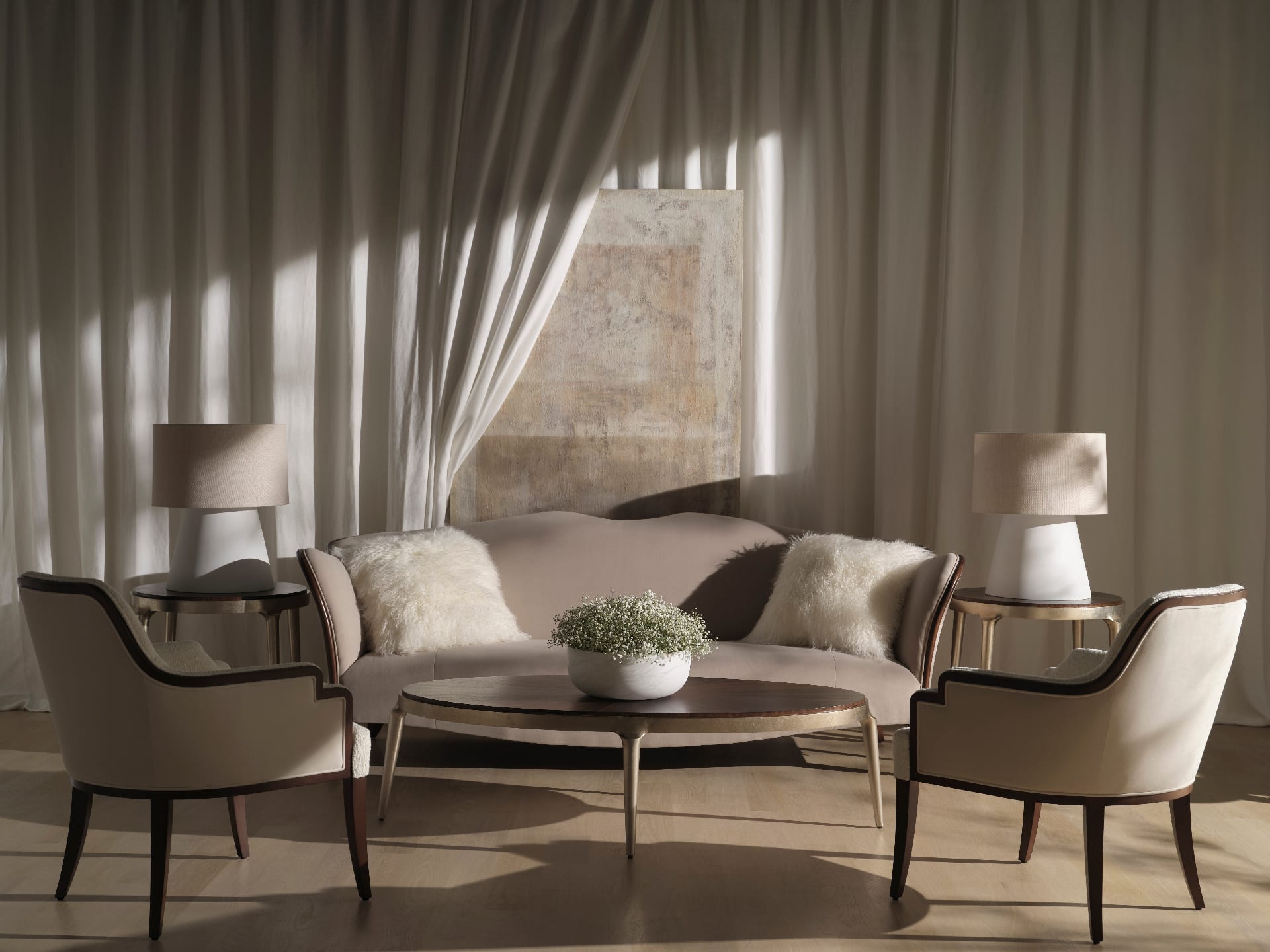 A serene sitting room in late afternoon light — a curved cream sofa flanked by a pair of linen armchairs, brass-footed side tables and a low oval coffee table beneath sheer drapery