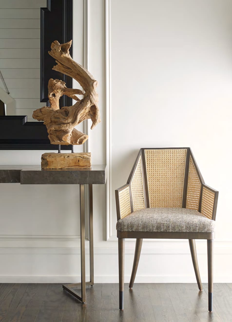An entry vignette — a caned occasional armchair beside a stone-topped console with a sculptural driftwood object and tall black-framed mirror