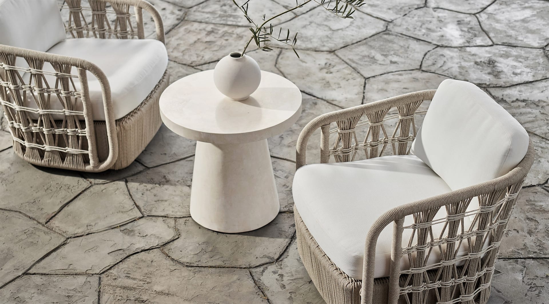 A flagstone terrace set with two open-lattice rope lounge chairs around a small cast-plaster side table, an olive sprig in a stone vessel