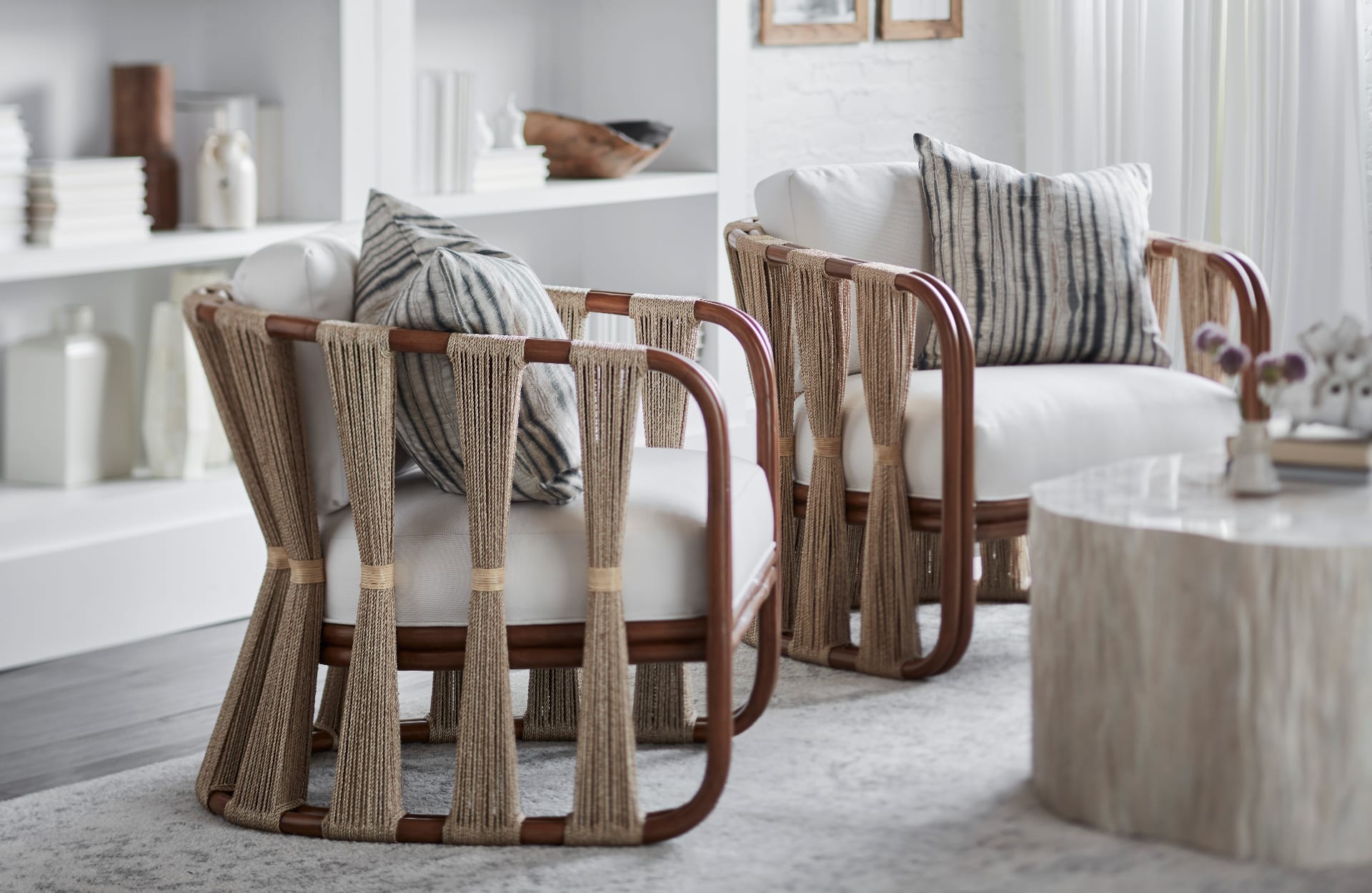 A pair of rope-strung rattan lounge chairs with ivory cushions and shibori-striped pillows, set on a soft grey rug beside a travertine drum table in a bright plaster-walled room