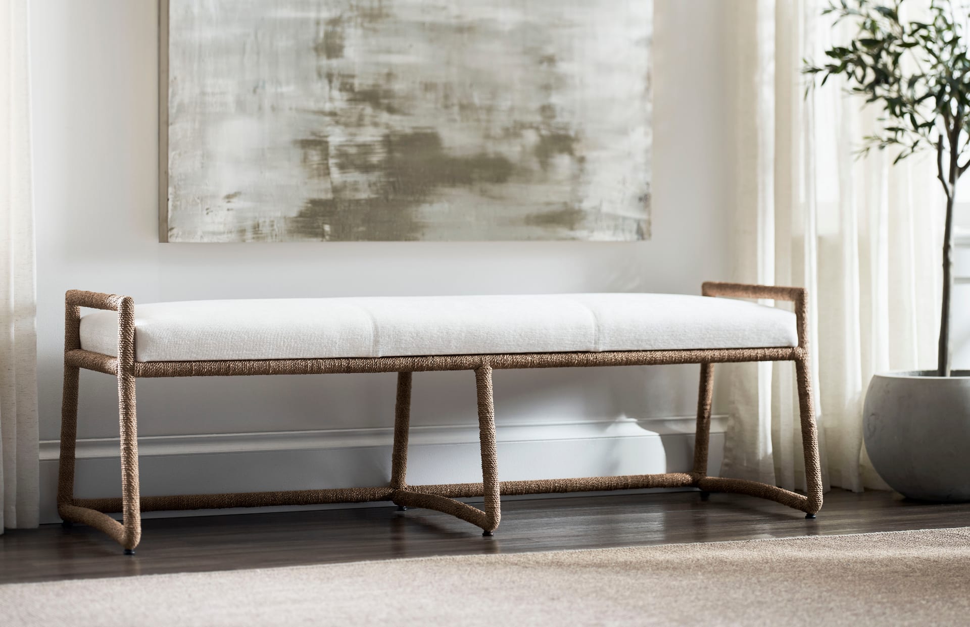 A long hand-wrapped natural rope bench with an ivory linen cushion, set beneath a soft abstract canvas in a bright plaster-walled room with sheer linen curtains and a potted olive tree