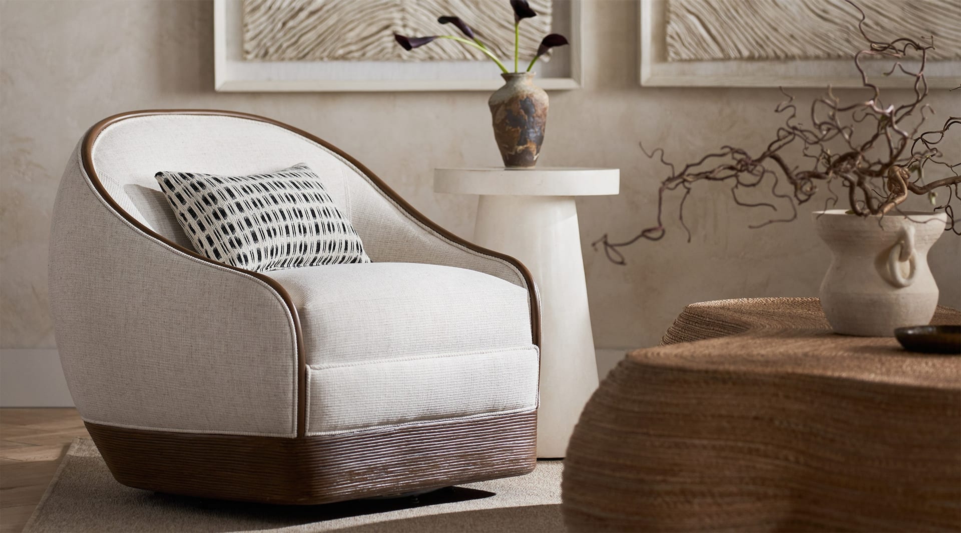 A linen-upholstered swivel chair on a reeded rattan plinth, set against a plaster wall and woven jute pouf in warm afternoon light