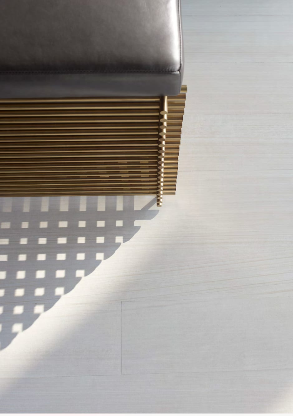 A leather-topped bench on a stacked brass rod base, raked light casting a graphic grid shadow across pale oak