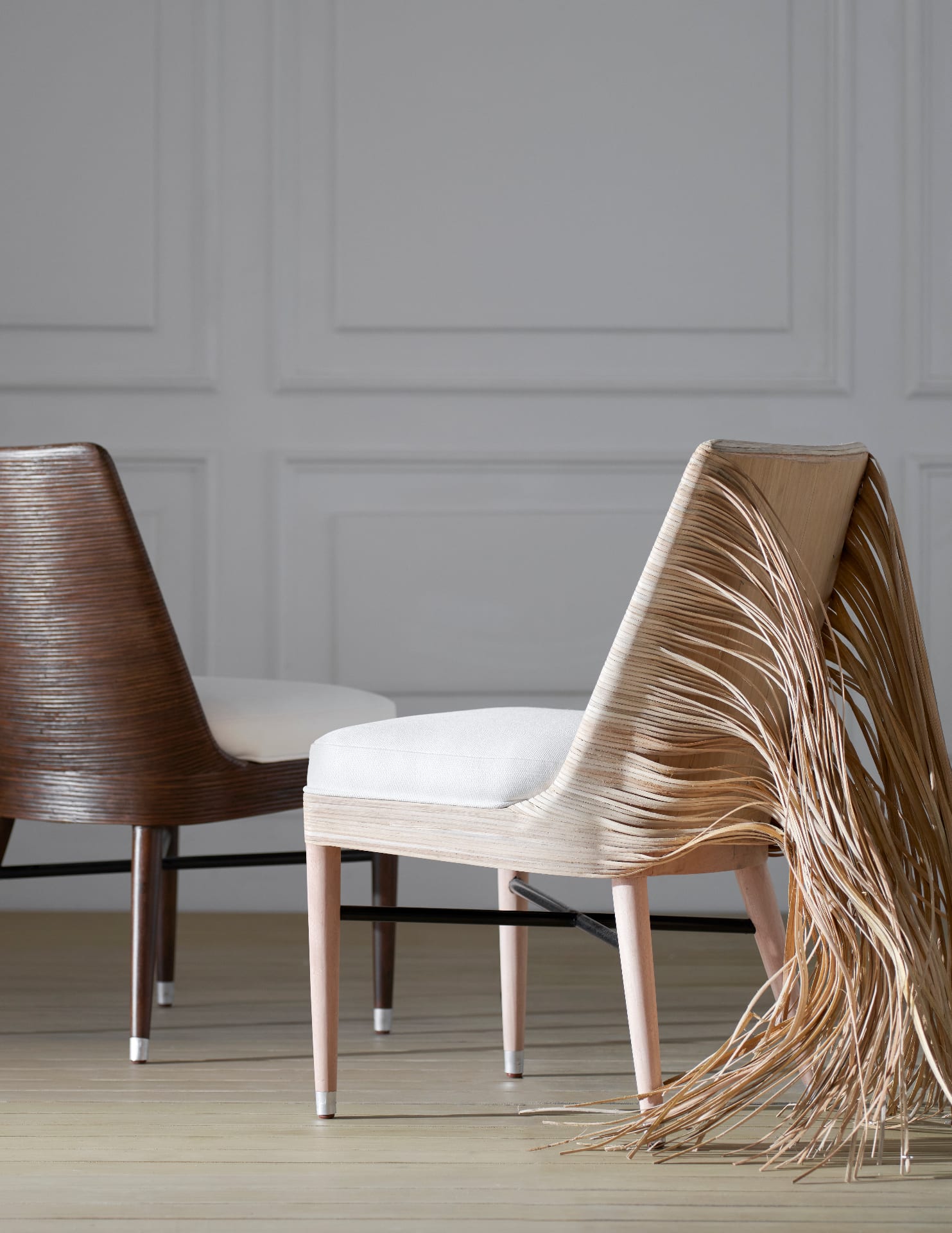 Two sculptural rattan-shell side chairs in profile against a panelled wall — one finished in deep walnut, the other left unfinished with long pale rattan strands trailing onto the floor
