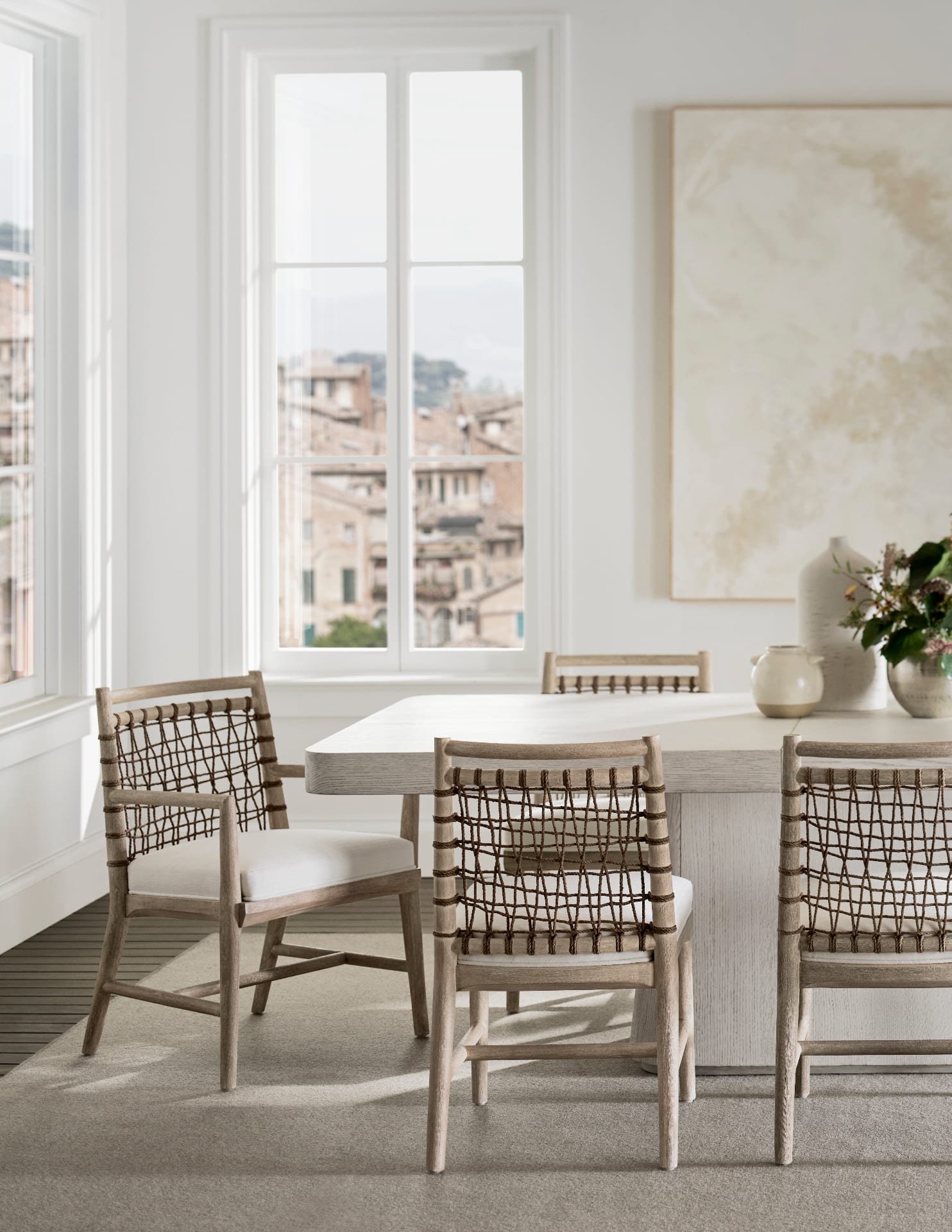 Cerused white oak dining chairs with hand-woven open-lattice rope backs and ivory linen seats, set around a pale oak table beside a tall window opening to a soft hilltop view