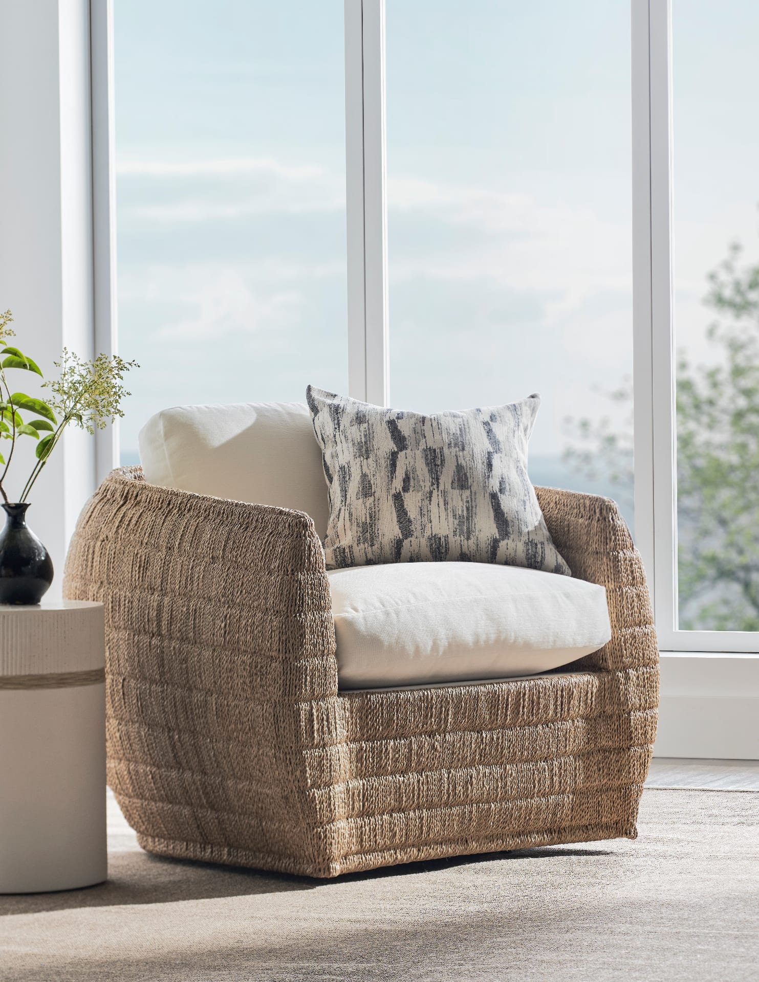 A deep, hand-woven seagrass swivel chair with a soft ivory linen cushion, set beside floor-to-ceiling windows looking out to a hazy blue sky