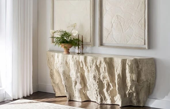 A sculptural cast-stone console with a craggy, rock-hewn front in pale ivory, set against a panelled plaster wall beneath two textured framed artworks, dressed with anthurium and a fern in a footed vessel